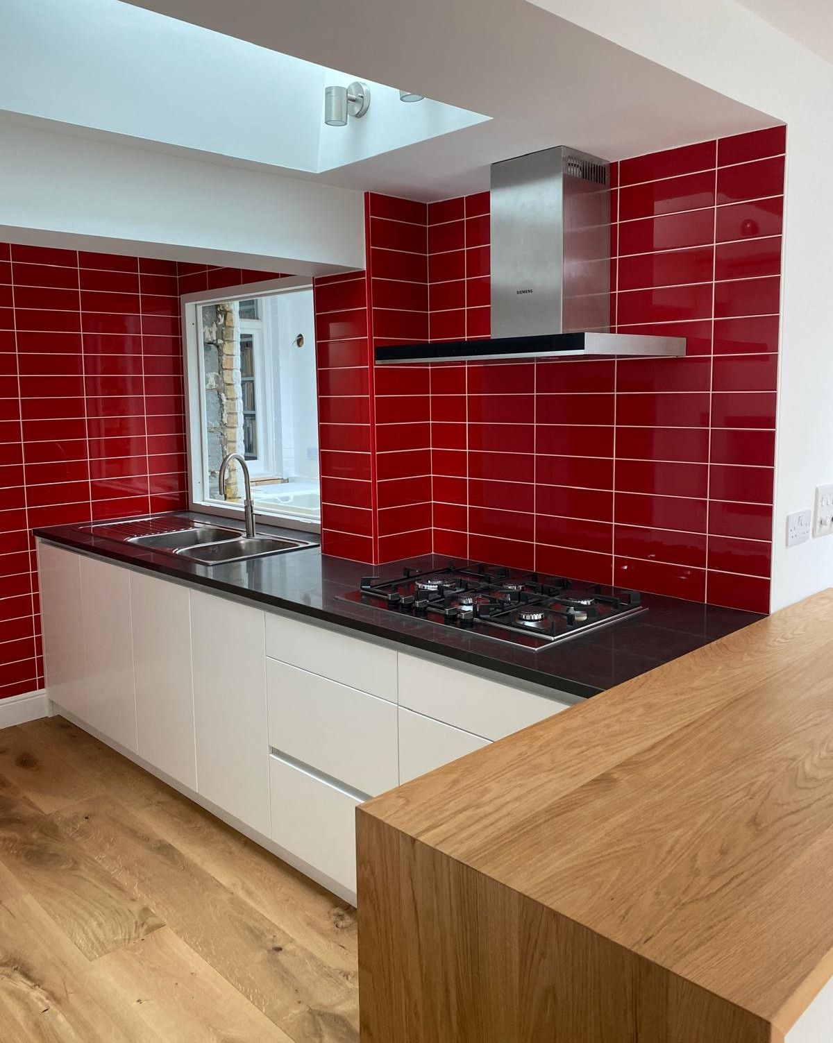 White kitchen island with black worktop, gas hob and glossy red tiled splashback under a skylight