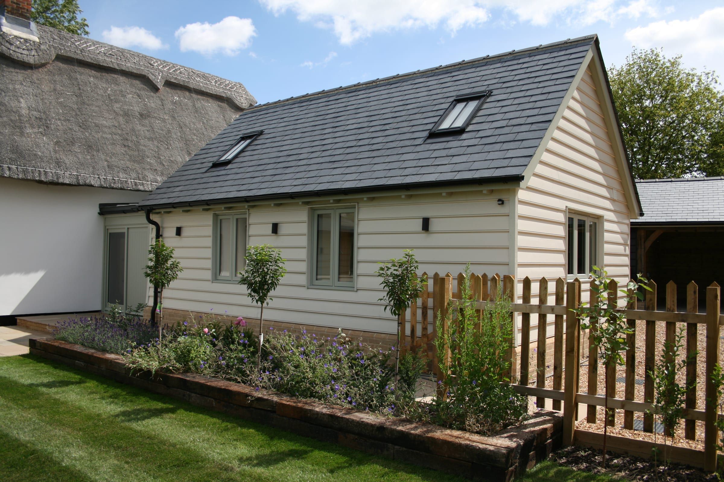 Timber-clad garden room annex with sage-green casement windows beside thatched cottage