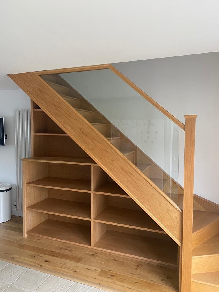 Understairs storage unit with glass balustrade panel above open oak shelving