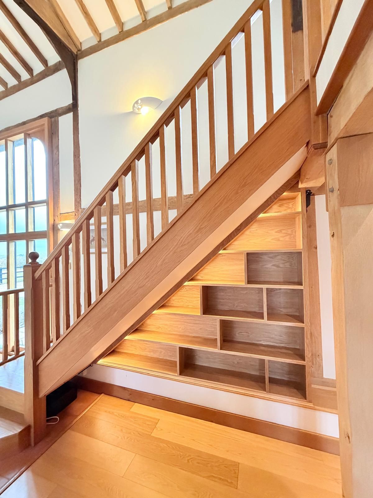 Angled oak understairs shelving with integrated lighting beside glazed hallway door